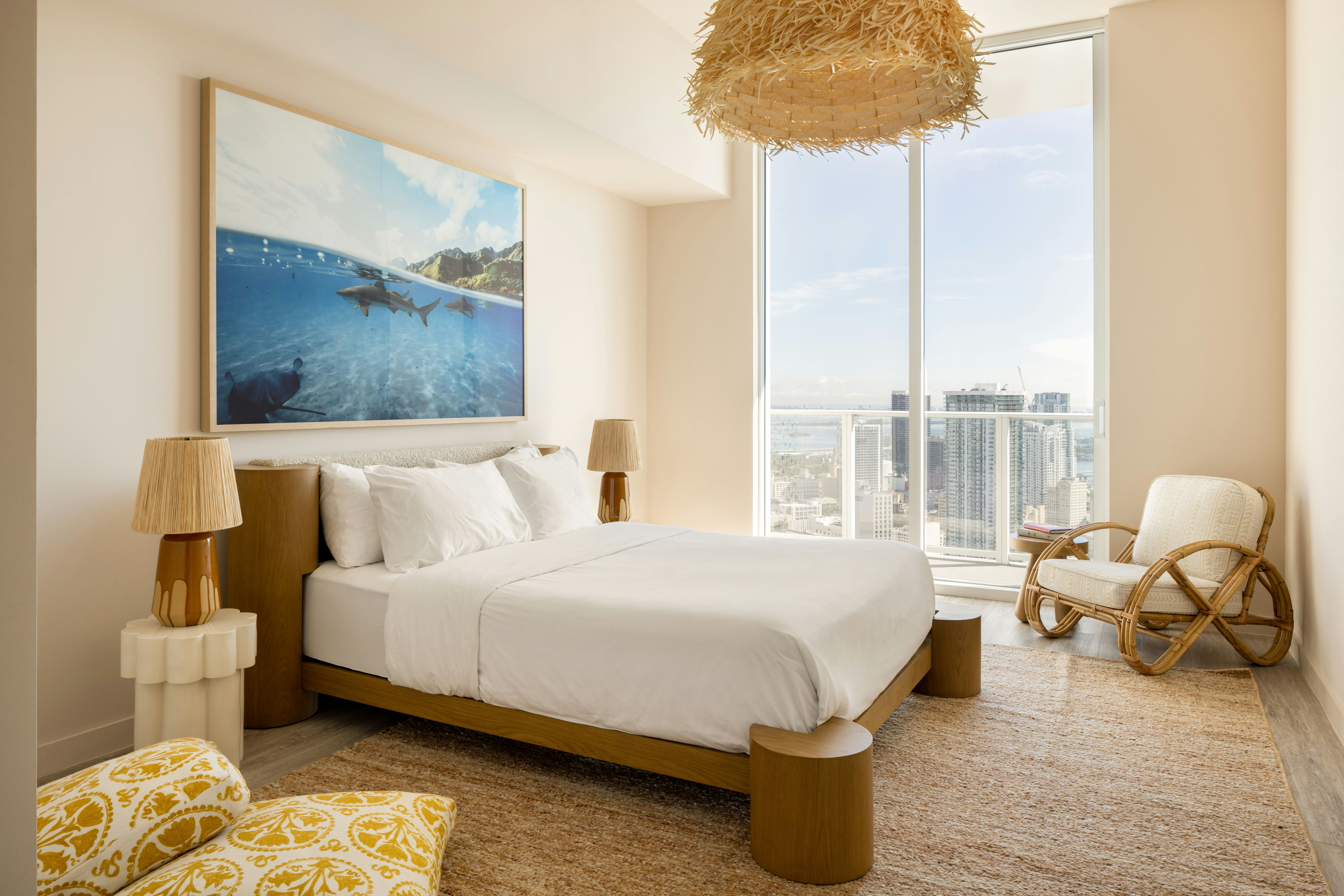 A large apartment bedroom with modern nightstands and lamps on each side, a comfortable chair, and a water painting above the headboard